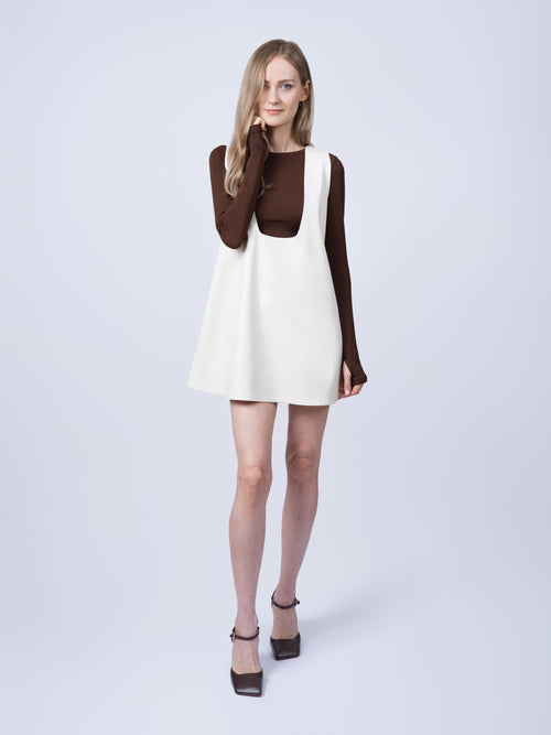 Women’s ecru vegan leather pinafore dress featuring a relaxed oversized silhouette and contemporary design. Front view styled with a fitted long-sleeve top, showcasing the clean lines and comfortable fit.
